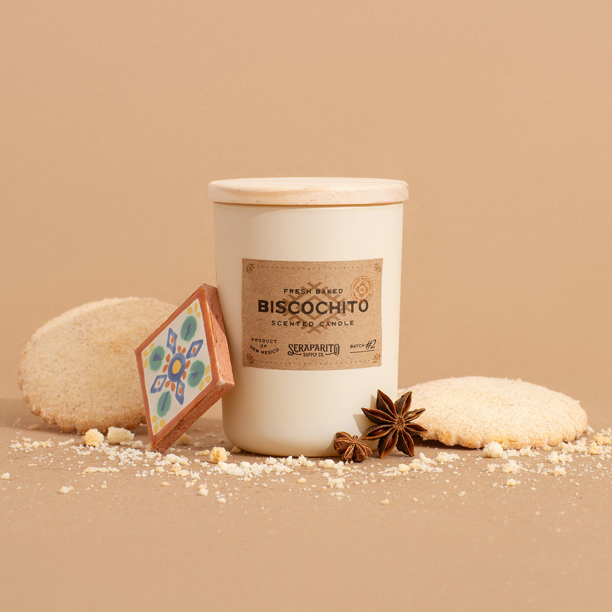 A biscochito scented candle in a tan glass jar with a label reading 'BISCOCHITO'. The candle is surrounded by what appears to be broken cookie pieces and whole cookies, with a cinnamon stick and anise star placed beside it, all against a beige background.